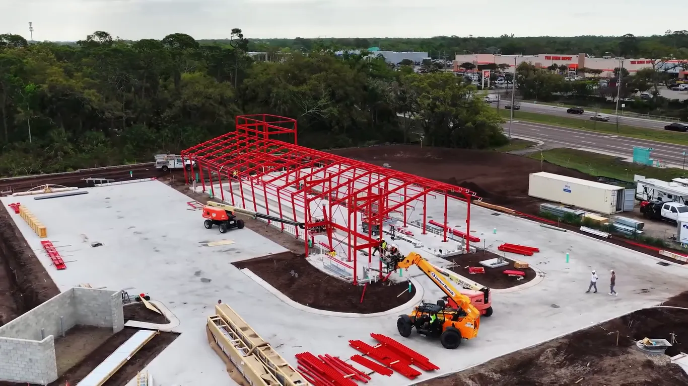 Aerial view of a new express car wash under construction: a red steel frame on a concrete slab, with a telehandler and stacks of steel beside it
