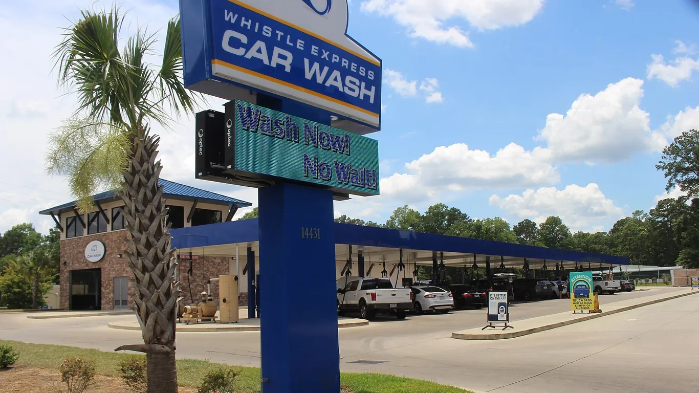 A Whistle Express car wash in Thomasville, Georgia: a tall sign reading Wash Now! No Wait!, a tunnel building and a long row of covered vacuum bays with cars parked in them