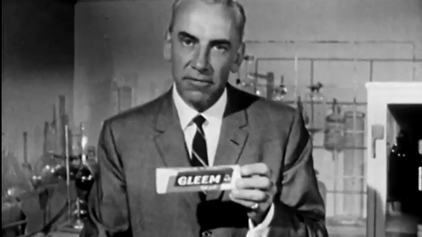A man in a suit holds up a tube of toothpaste in an old black and white television advertisement
